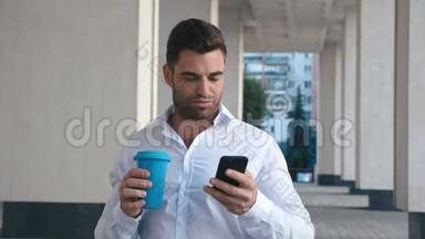 年轻人在办公大楼附近的城市用智能<strong>手机短信</strong>。 英俊的年轻商人喝咖啡